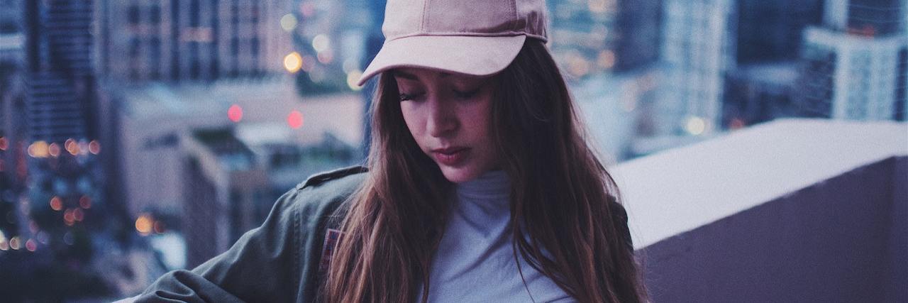 Why Feeling Too Much Feels Lonely A woman wearing a hat looking down, standing on a roof that overlooks city skyscrapers
