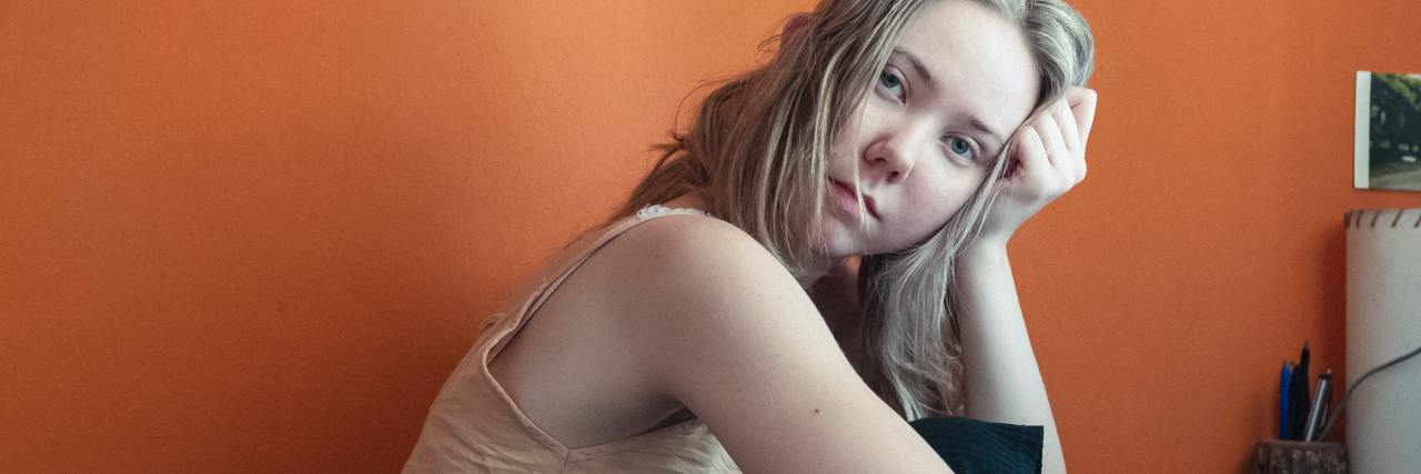 photo of woman resting her head on her hand at computer desk, looking sideways into camera, framed by an orange wall