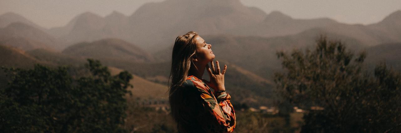 The Wim Hof Method Can Help You Breathe During COVID-19 photo of a woman standing outside with mountains in background, her head raised and eyes closed, breathing deeply