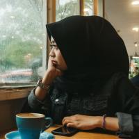 Woman wearing a hijab looks out a window into a rainy landscape with a cup of coffee in front of her