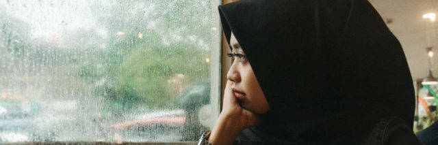 Woman wearing a hijab looks out a window into a rainy landscape with a cup of coffee in front of her