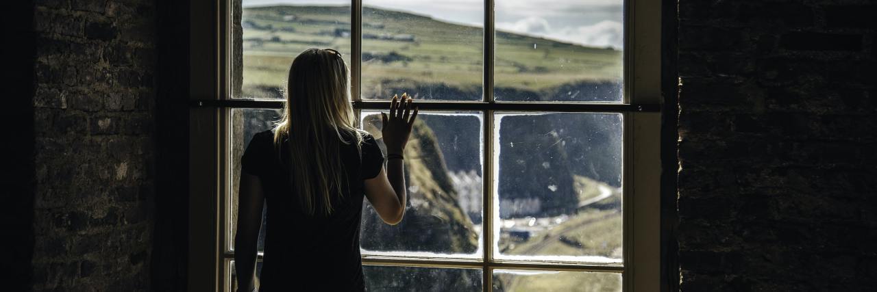 Asking for Help Is Hard, but Dealing With Depression Alone Is Worse photo of woman looking out of tall window at cliff landscape, raising her hand to touch the glass, with her back to the camera