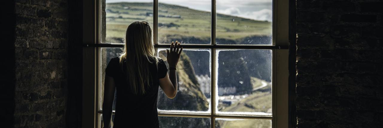 photo of woman looking out of tall window at cliff landscape, raising her hand to touch the glass, with her back to the camera