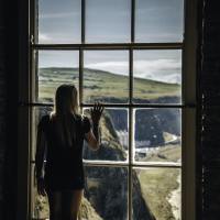 photo of woman looking out of tall window at cliff landscape, raising her hand to touch the glass, with her back to the camera