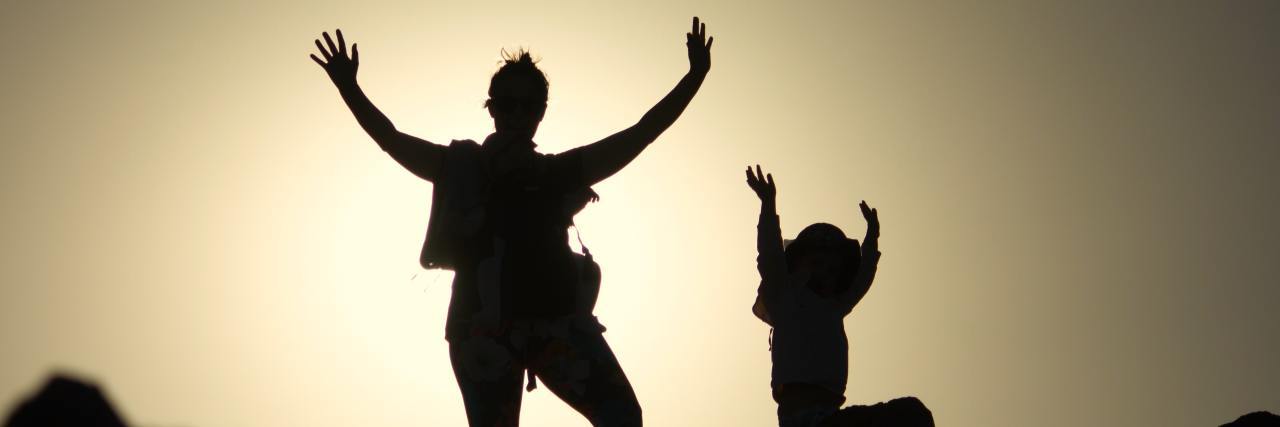 photo of mother and child having fun with arms raised, silhouetted against featureless sky at sunset