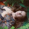 photo of woman with her hands crossed on her chest, lying on the ground in a forest surrounded by ferns and looking into camera
