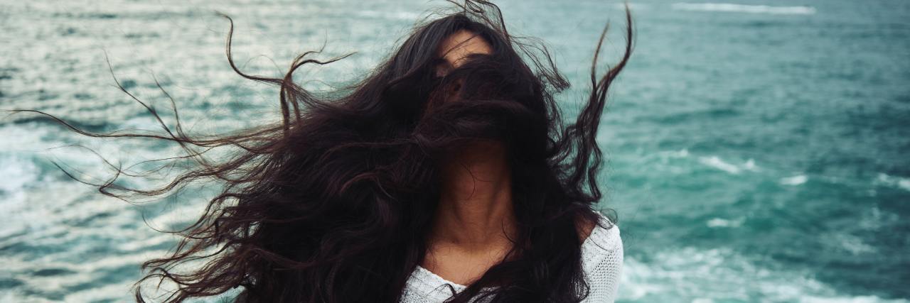 Struggling to Accept My New Reality of Life With Chronic Illness woman with dark hair flying around her face with the ocean in the background