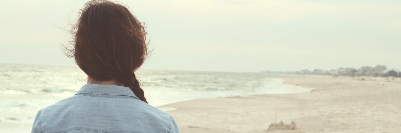 A young white woman with her back to the camera, standing on a beach looking at the ocean