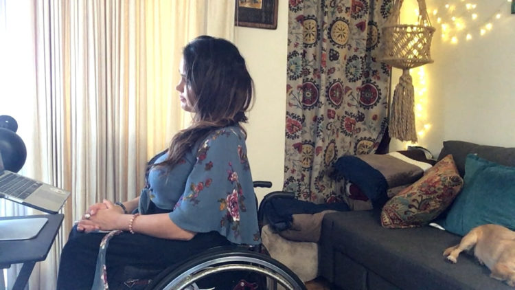 Actress Santina Muha, a woman with long brown hair wearing a blue floral print shirt and sitting in her wheelchair facing a computer screen