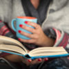 Woman reading a book and holding a mug of hot beverage.