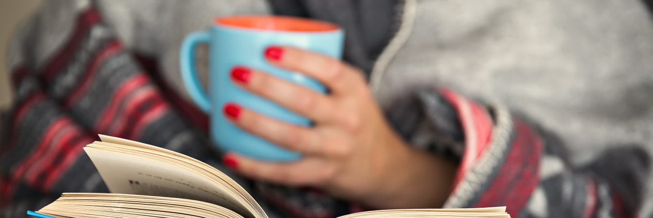 Woman reading a book and holding a mug of hot beverage.