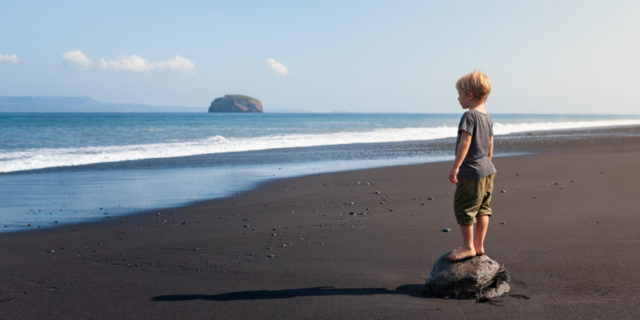 Child standing on big stone on black sand sea beach