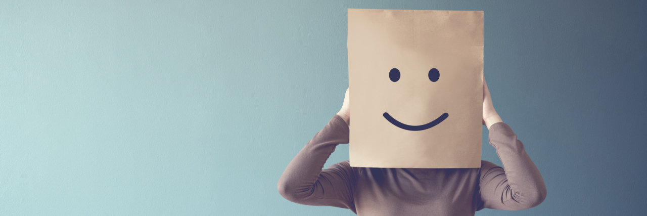 A white woman in a sweater holding a brown paper bag over her head with a smiley face on it.