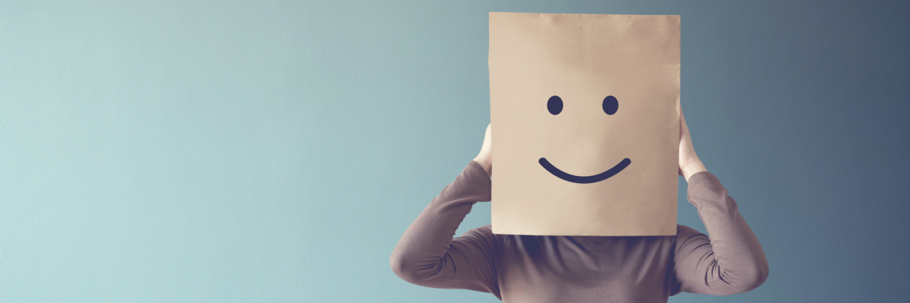 Positive Thinking Will Not 'Cure' My OCD A white woman in a sweater holding a brown paper bag over her head with a smiley face on it.