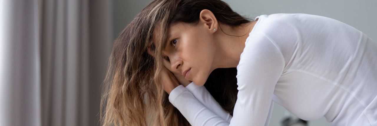 Why I Always Think About My Chronic Illnesses a woman looking outside from her bedroom with hand on forehead