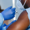hand of medical staff in blue glove injecting coronavirus covid-19 vaccine in vaccine syringe to arm muscle of man wearing scrubs