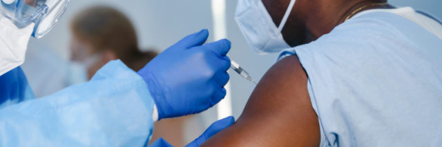 hand of medical staff in blue glove injecting coronavirus covid-19 vaccine in vaccine syringe to arm muscle of man wearing scrubs