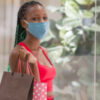 photo of a young woman in a face mask, looking into the camera in front of a shopping mall window and carrying shopping bags