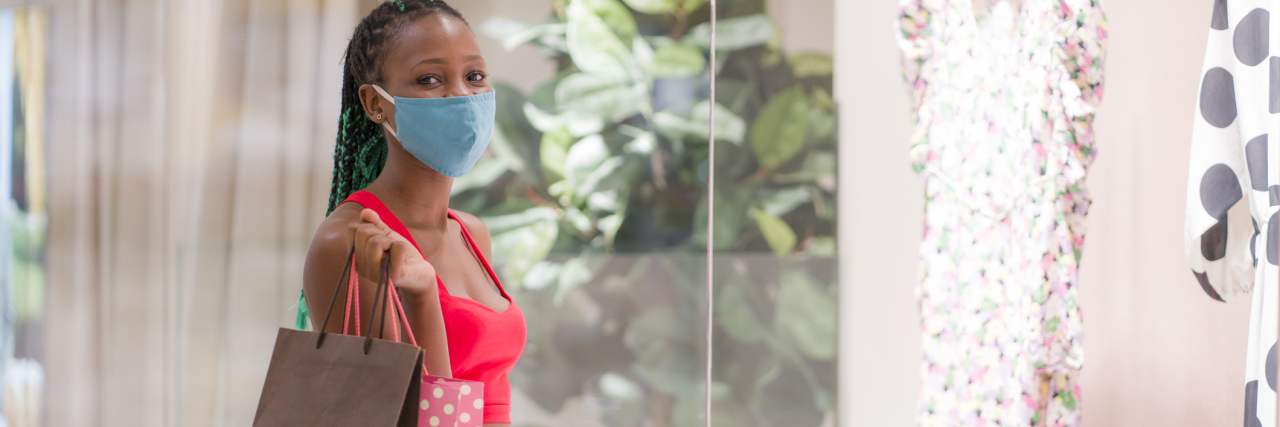 Compulsive Shopping When I'm Struggling With Depression photo of a young woman in a face mask, looking into the camera in front of a shopping mall window and carrying shopping bags