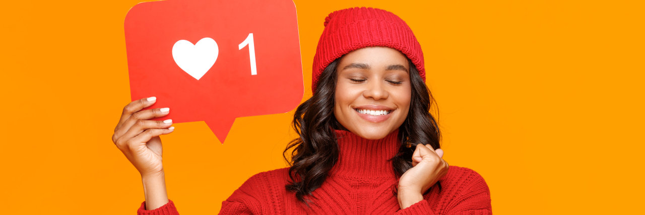 Should You Post About Getting the COVID-19 Vaccine on Social Media? Woman wearing a red sweater and red beanie holding up a posted on a heart/like button from social media