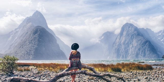 Woman sitting on driftwood lakeshore against snowcapped mountains in New Zealand