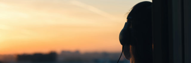 back of a girl leaning against the side of a window with headphones on at sunset
