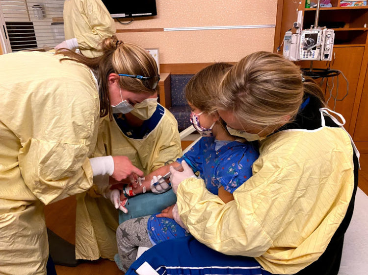 Contributor's daughter with her arm out while surrounded by three nurses putting an IV in it 