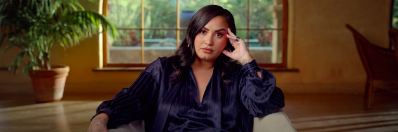 Demi Lovato Reveals Why Overdose Left Lasting Brain Damage Demo Lovato site on a couch wearing a dark blue jumpsuit