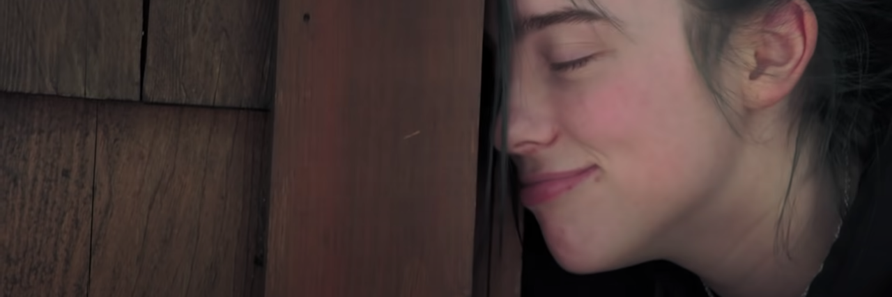 Mental Health Review of Billie Eilish: The World's A Little Blurry screenshot from Billie Eilish documentary showing her leading her forehead against door frame, eyes closed and smiling