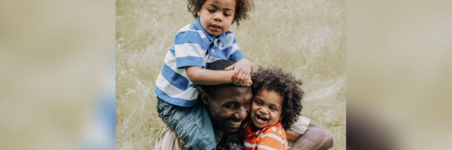 photo of a Black father and two children, one in his arms and the other on his shoulder, laughing and smiling together