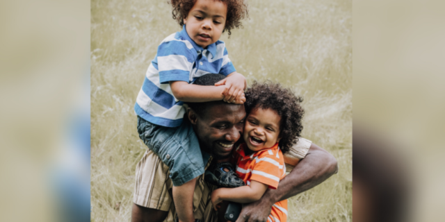 photo of a Black father and two children, one in his arms and the other on his shoulder, laughing and smiling together