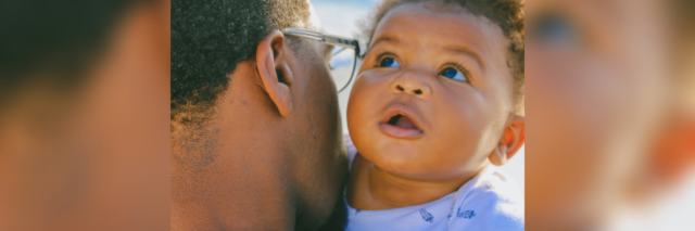 close up photo of Black father with young baby over his shoulder, sea in background