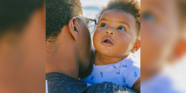 Black History Month: Reflections on Black Trauma and My Biracial Trans Identity close up photo of Black father with young baby over his shoulder, sea in background