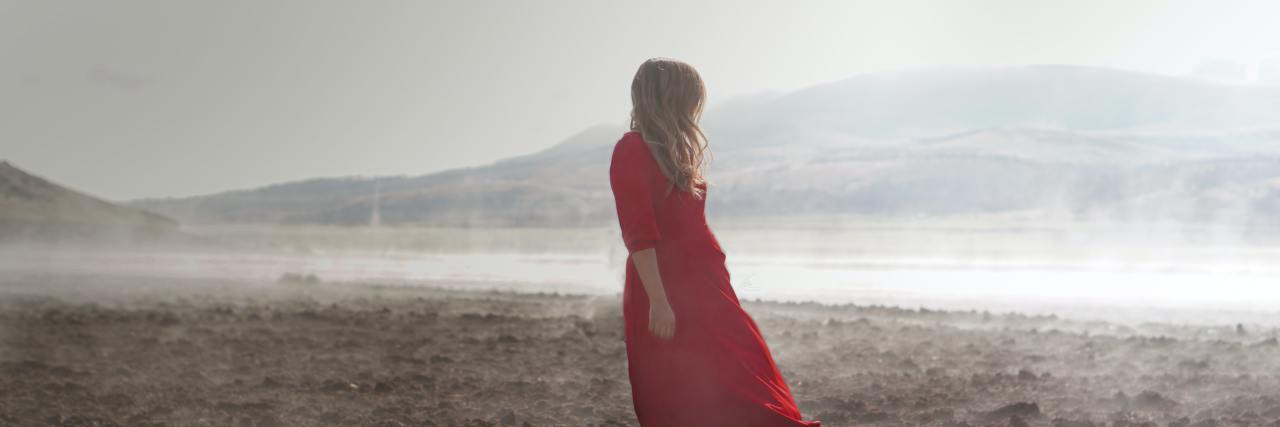 A blond white woman in a red dress looking over her shoulder at the ocean, standing on a beach