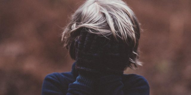 How My Childhood Loneliness Grew Into Anxiety photo of child with blonde hair, wearing gloves and covering face and eyes, posing in front of a blurry forest background
