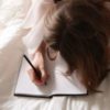 photo of woman lying on bed with white sheets, writing in journal