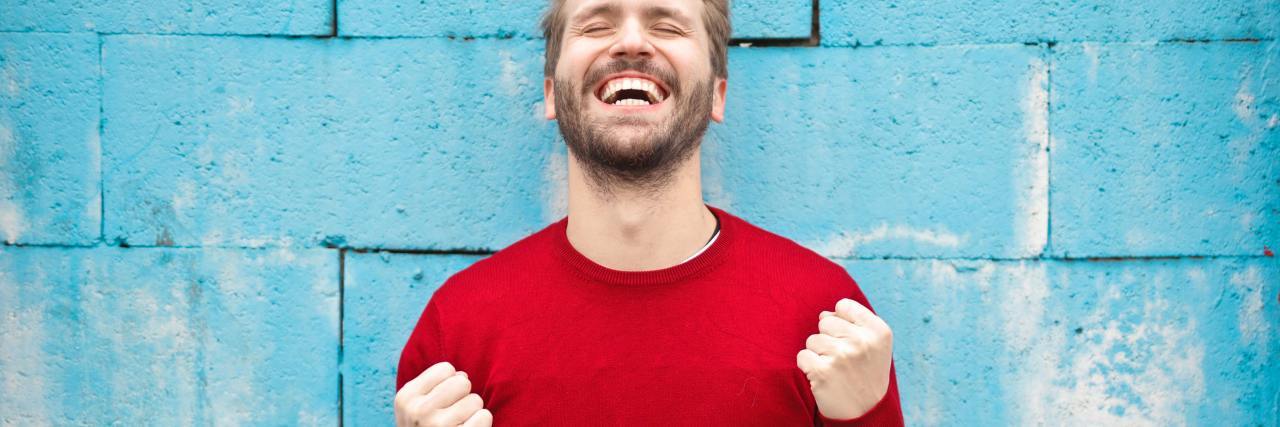Let Go of What People Think of You When You Have Schizophrenia photo of man wearing a red shirt standing against blue brick wall, fists raised in celebration of success