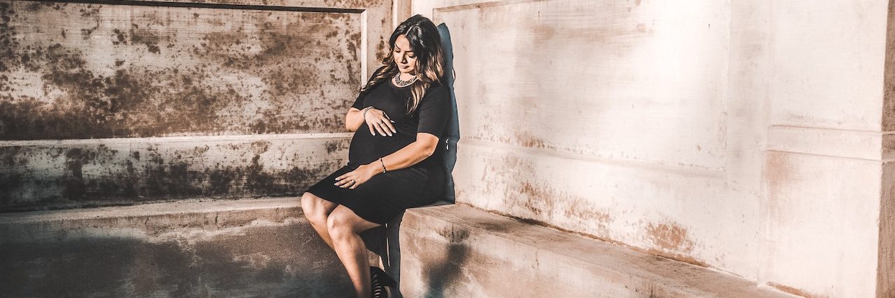What You Should Know About Perinatal Depression A pregnant woman wearing a black dress sits in the corner of an empty gray room