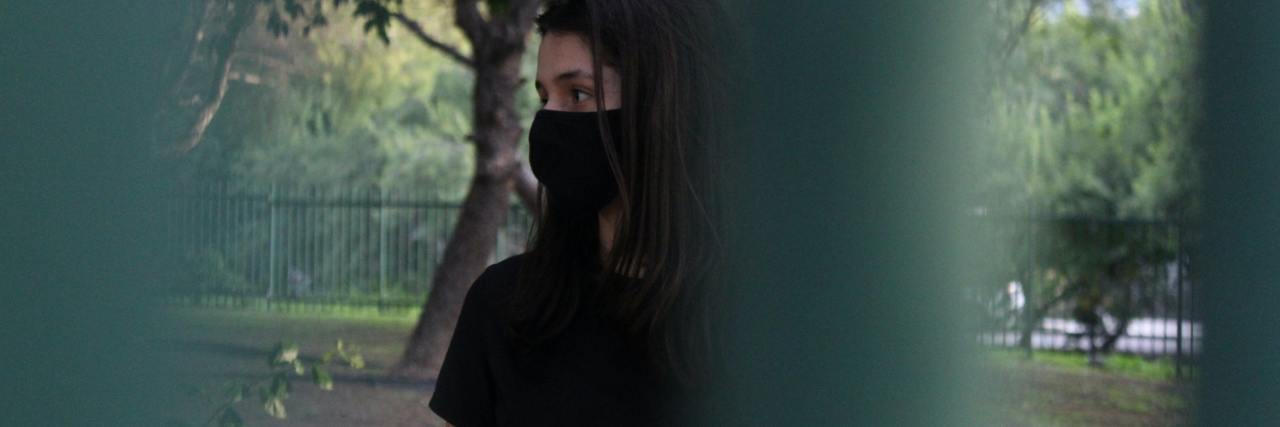 photo of woman behind fence, standing in a park with a tree behind her, wearing a black face mask and looking scared