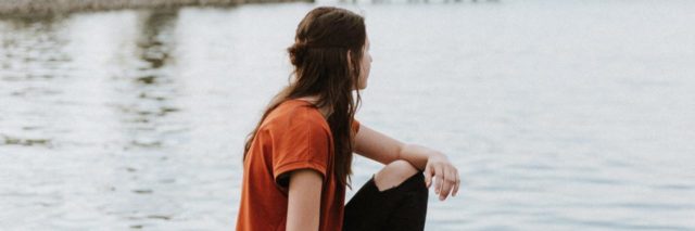 photo of woman sitting on a rock by some water, a pier in the background, looking off into distance