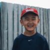 Young boy wearing a red baseball cap and navy sweatshirt smiles in front of a wooden fence