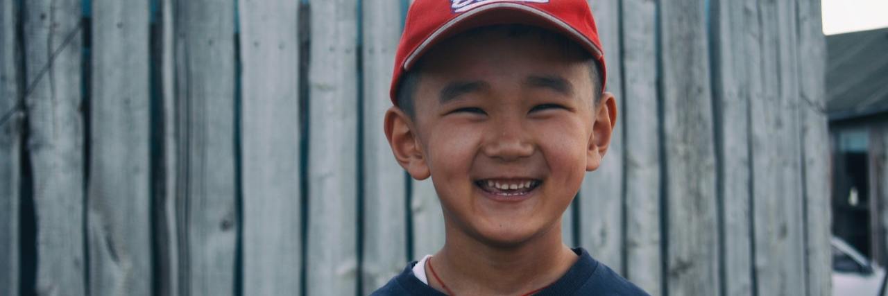 Young boy wearing a red baseball cap and navy sweatshirt smiles in front of a wooden fence