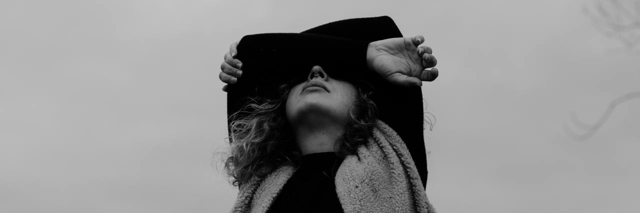 black and white photo of woman covering her eyes with her arms, taken from below with a backdrop of cloudy sky