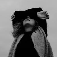 black and white photo of woman covering her eyes with her arms, taken from below with a backdrop of cloudy sky