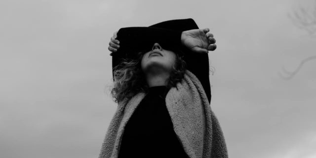 black and white photo of woman covering her eyes with her arms, taken from below with a backdrop of cloudy sky