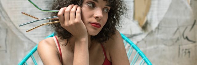 A woman with dark, curly hair wears a red tank top while sitting on a green chair holding several reusable straws in her hand
