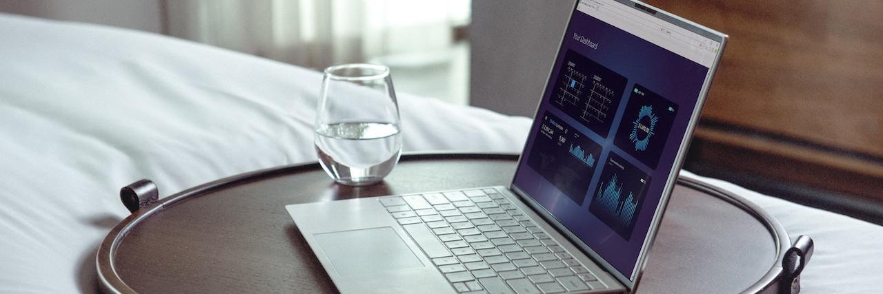 Remote Jobs for People With a Disability or Chronic Illness Open laptop alongside a glass of water on a wooden bed table