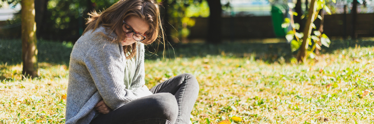 photo of a young woman sitting outdoors on grass, holding tummy in pain