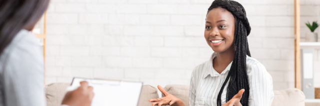 A young Black woman sitting on a therapist's couch smiling with her hands out