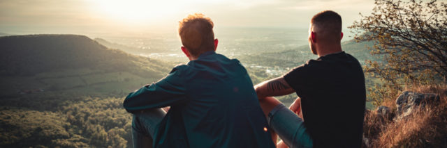 2 young men sitting on top of a mountain overlooking the sunset